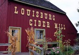 Red barn with words "Louisburg Cider Mill" on the side with bunches of wheat in front